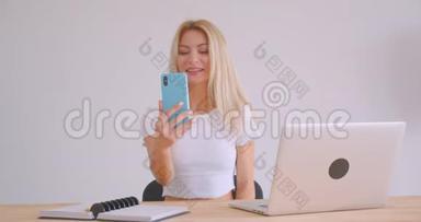 一位成年白种人金发女商人坐在笔记本电脑前笔记本电脑前，在手机上打了个视频电话
